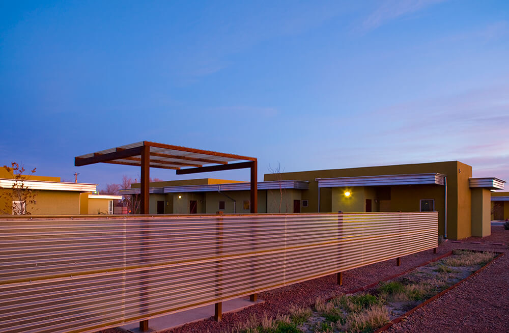 Rust-steel trellis and corrugated steel fence at dusk with lit single-story homes beyond at Wilshire Gardens