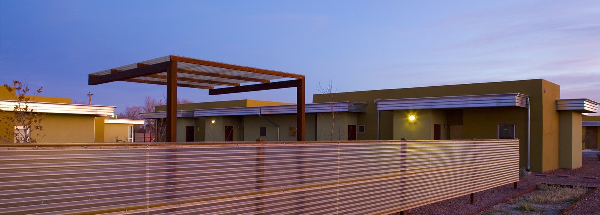 Wide dusk view of Wilshire Gardens in Roswell with single-story homes, a steel trellis, and warm entry lights along the fence line